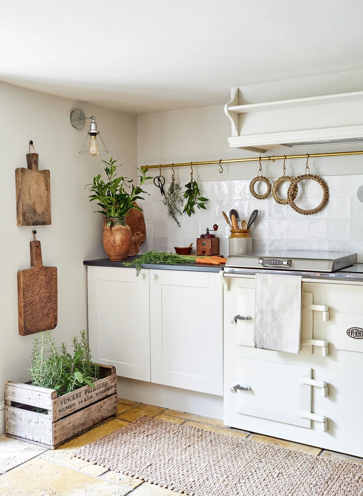 A white kitchen corner has a pale range cooker, simple framed cabinets, a wall rail with hooks, hanging wood boards and a dark counter.