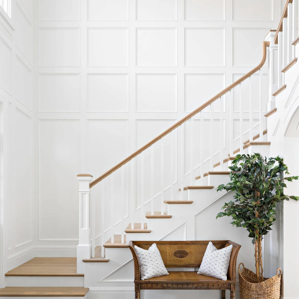 White rectangular wall paneling rises beside wood stairs, dark rails and a cane-backed bench.
