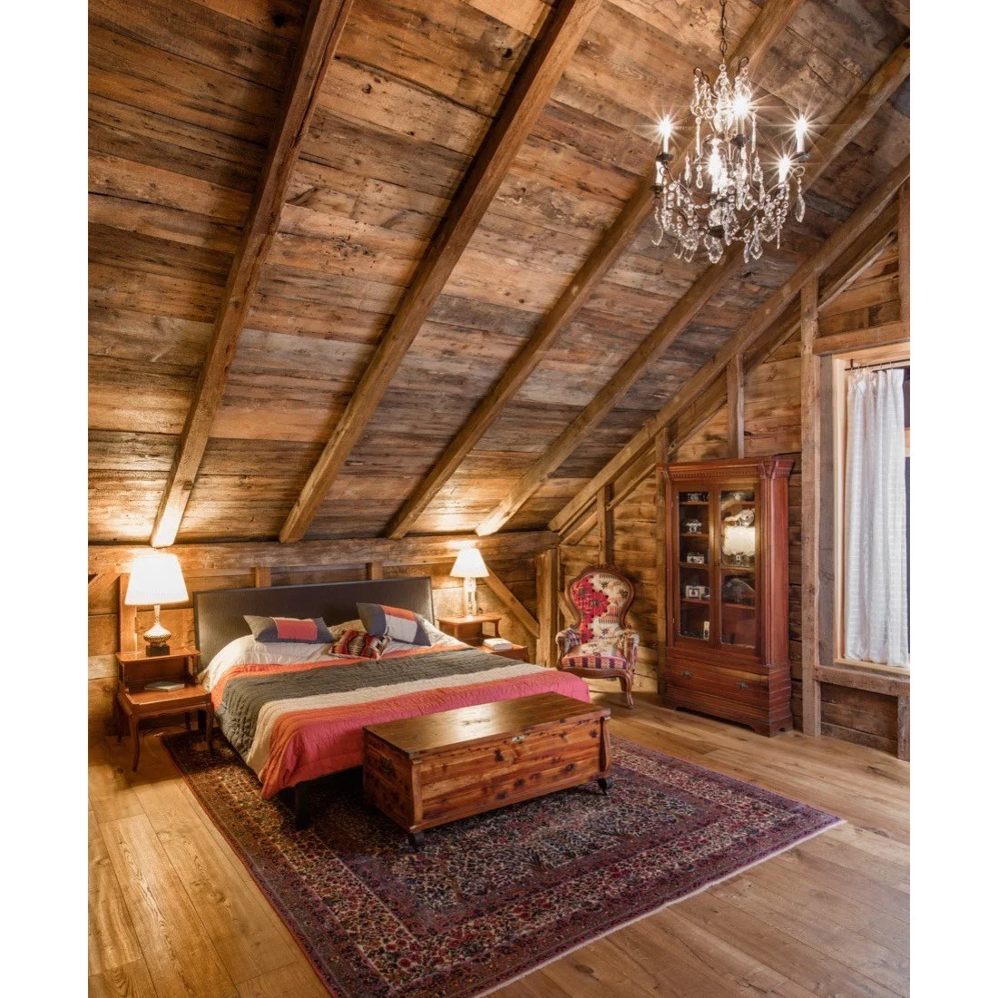 A bed with multicolored quilt beneath a sloping timber roof and glass chandelier beside a wood chest