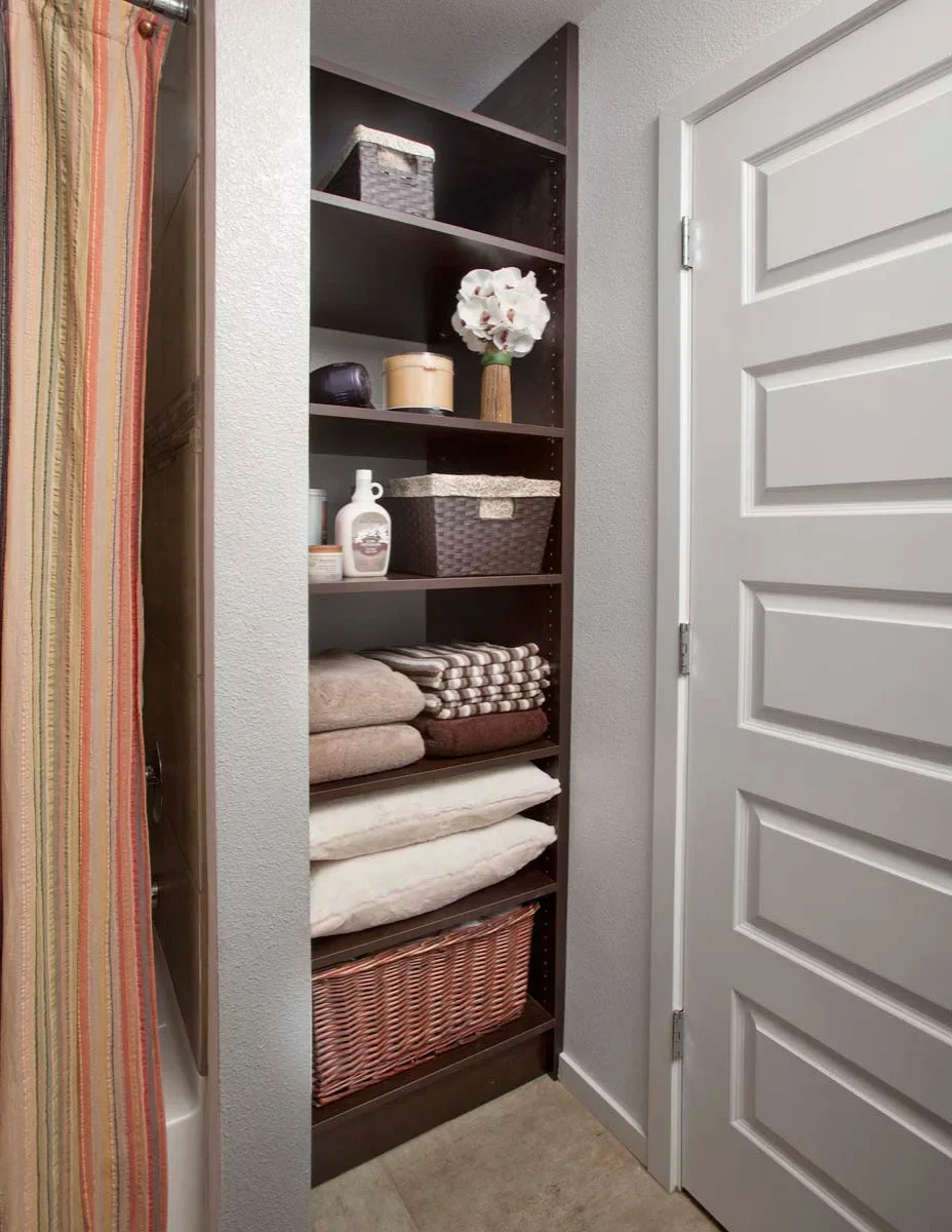 Slim dark linen cupboard with folded towels, pillows and a large woven basket above a carpeted floor