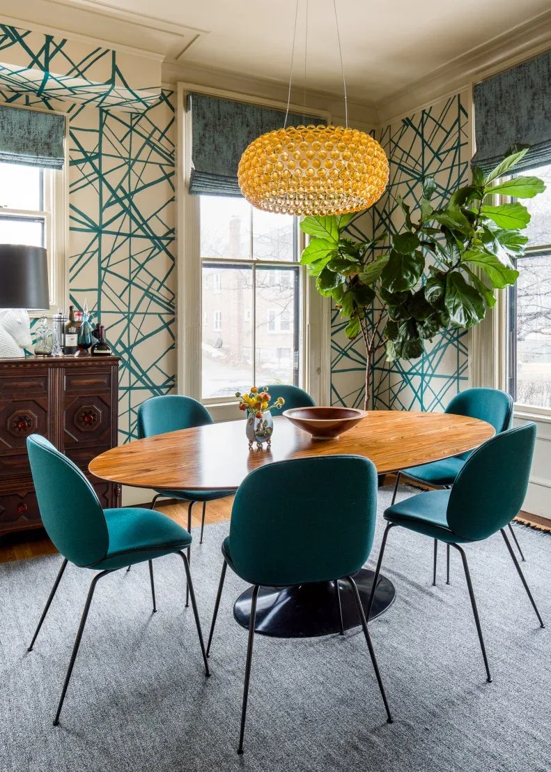 Teal angular wall pattern between dining windows behind an oval wood table and teal upholstered chairs