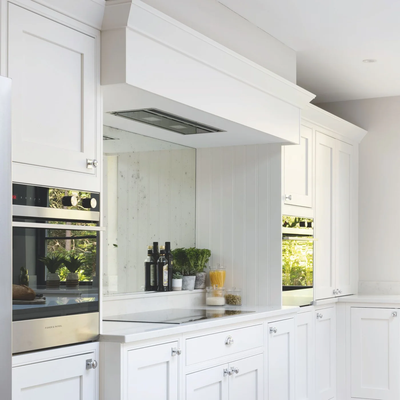 White framed cabinets and built-in ovens flank a cooktop beneath a broad white overmantel and mirrored backsplash.