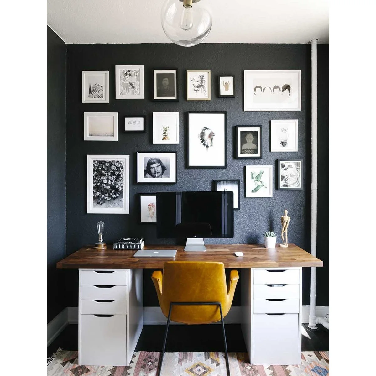 Wood-top desk between white drawer units with a yellow chair and gallery wall on a dark background