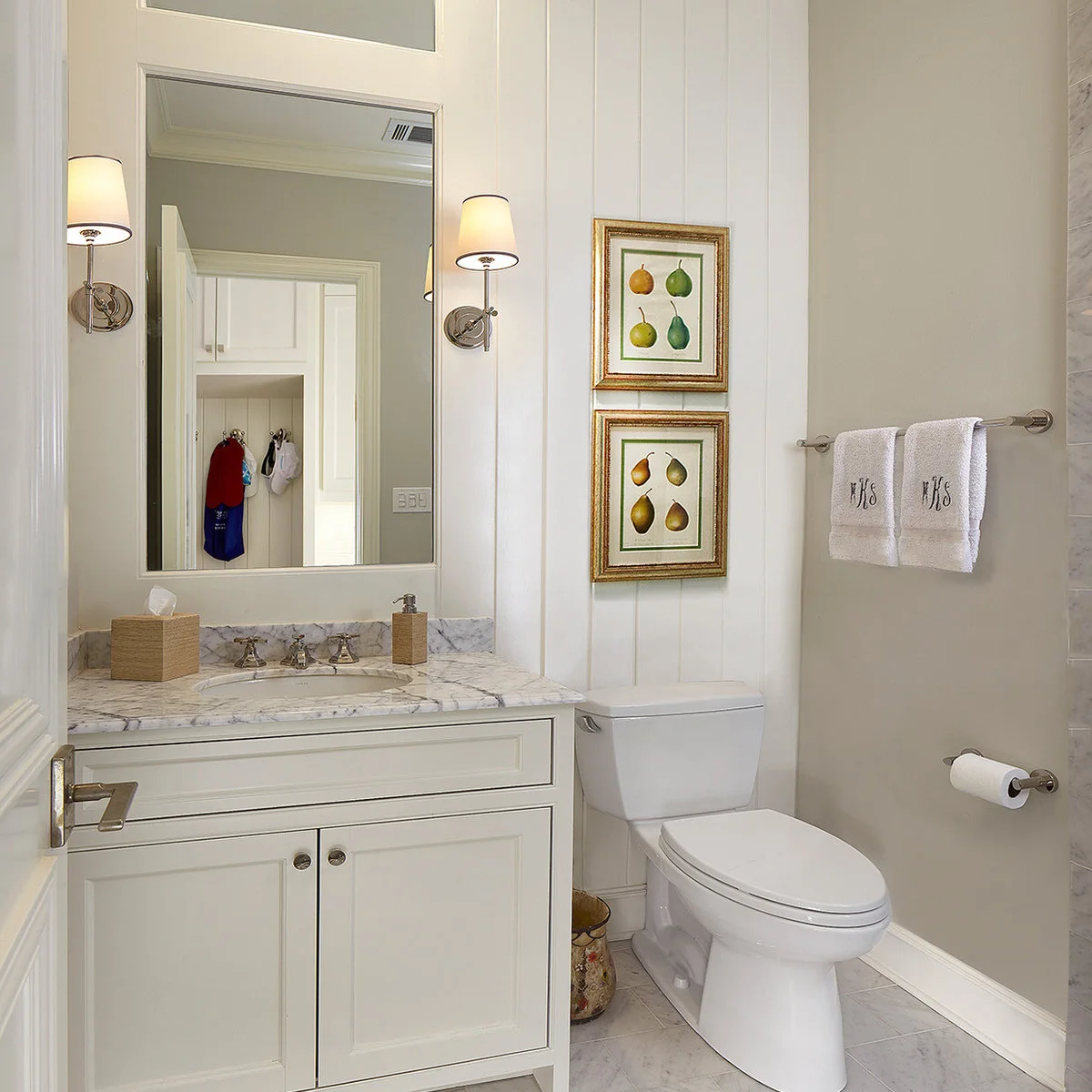 A pale powder room with a white vanity, paired fruit prints above the toilet and a long towel rail.