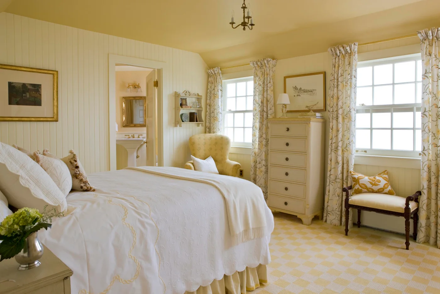 Cream-and-yellow bedroom with white bedding, floral curtains, a tall chest and two individual chairs