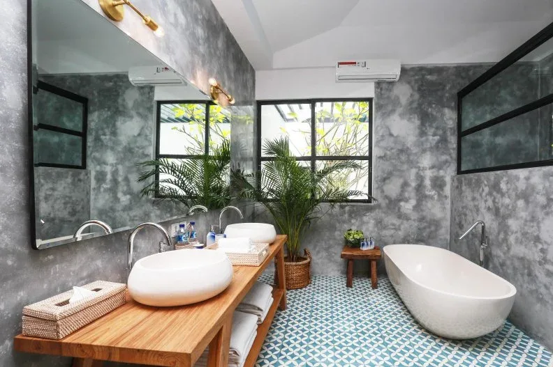 A timber counter with two white basins sits beside a large plant in a mottled gray bathroom.