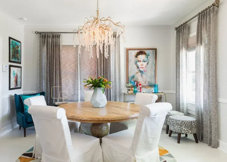 Round wooden table with pale covered chairs, a leopard-patterned stool and an ornate pink chandelier near windows.