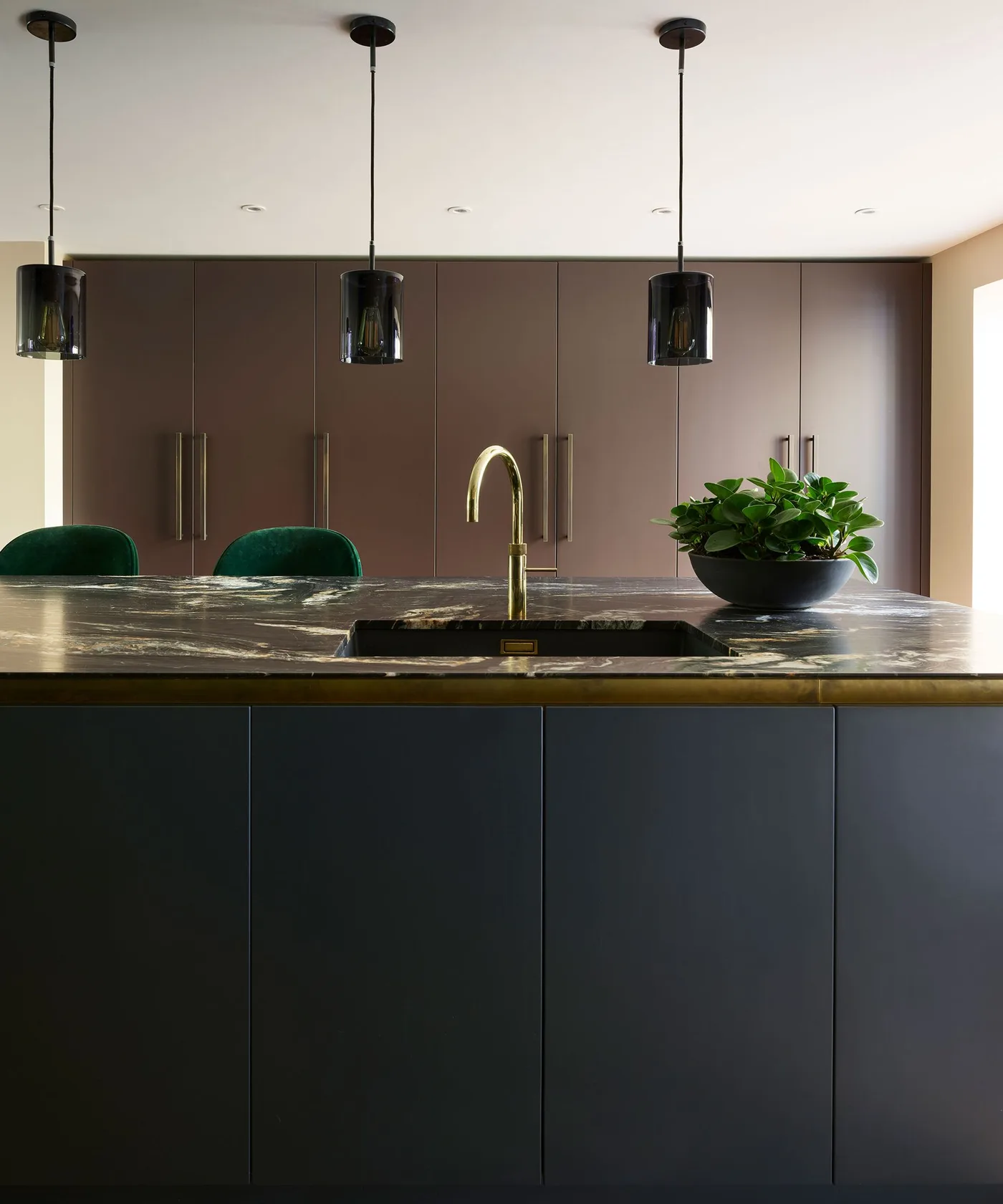 Kitchen with dark flat-front island, strongly veined dark counter, brown tall cabinets, long warm metal handles, three dark pendants, and green chairs.
