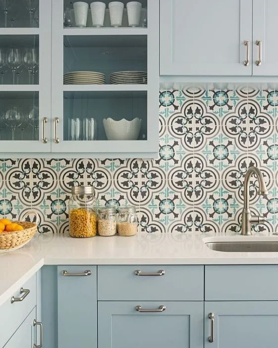 Pale blue cabinets and glass-front cupboards surround a sink beneath larger teal, black and pale patterned tiles with jars along the counter