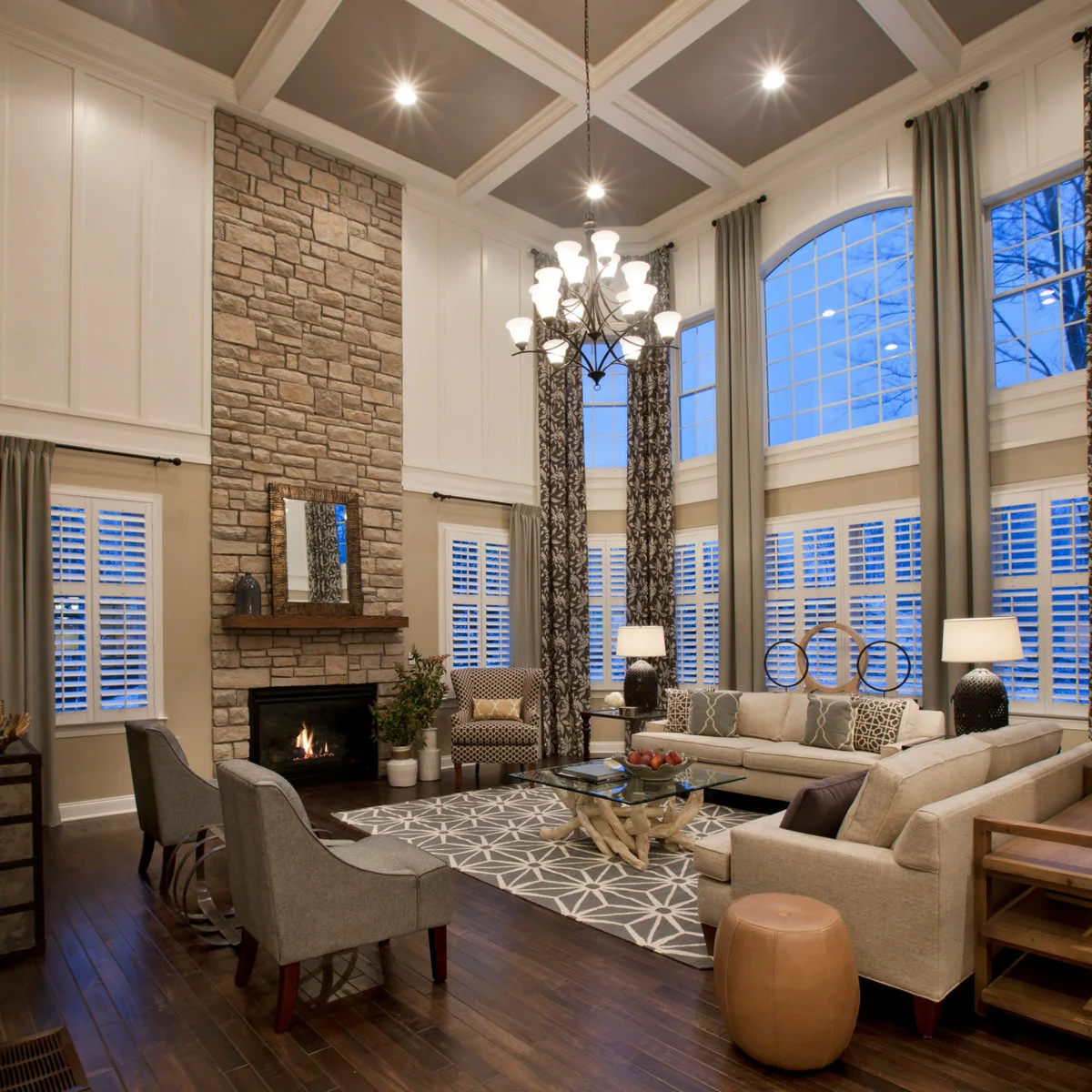 A double-height room with lower white shutters, tall curtain panels, pale seating and a stone fireplace beneath a coffered ceiling.
