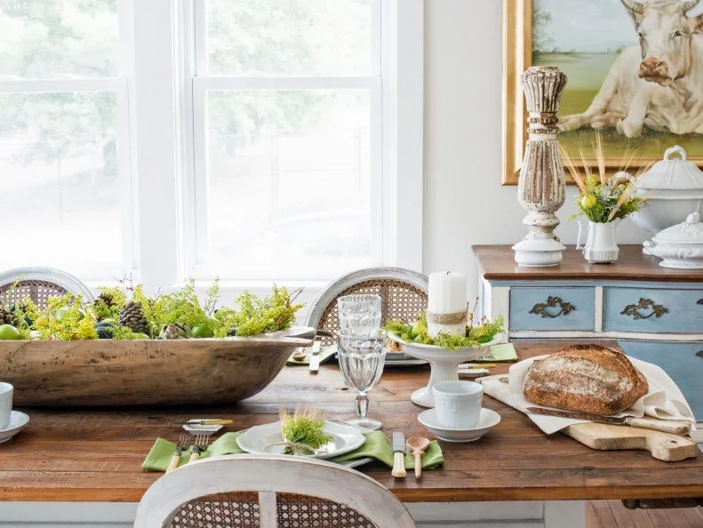 A shallow wooden bowl holds bright green foliage and pinecones beside green napkins, pale crockery and bread on a wooden dining table