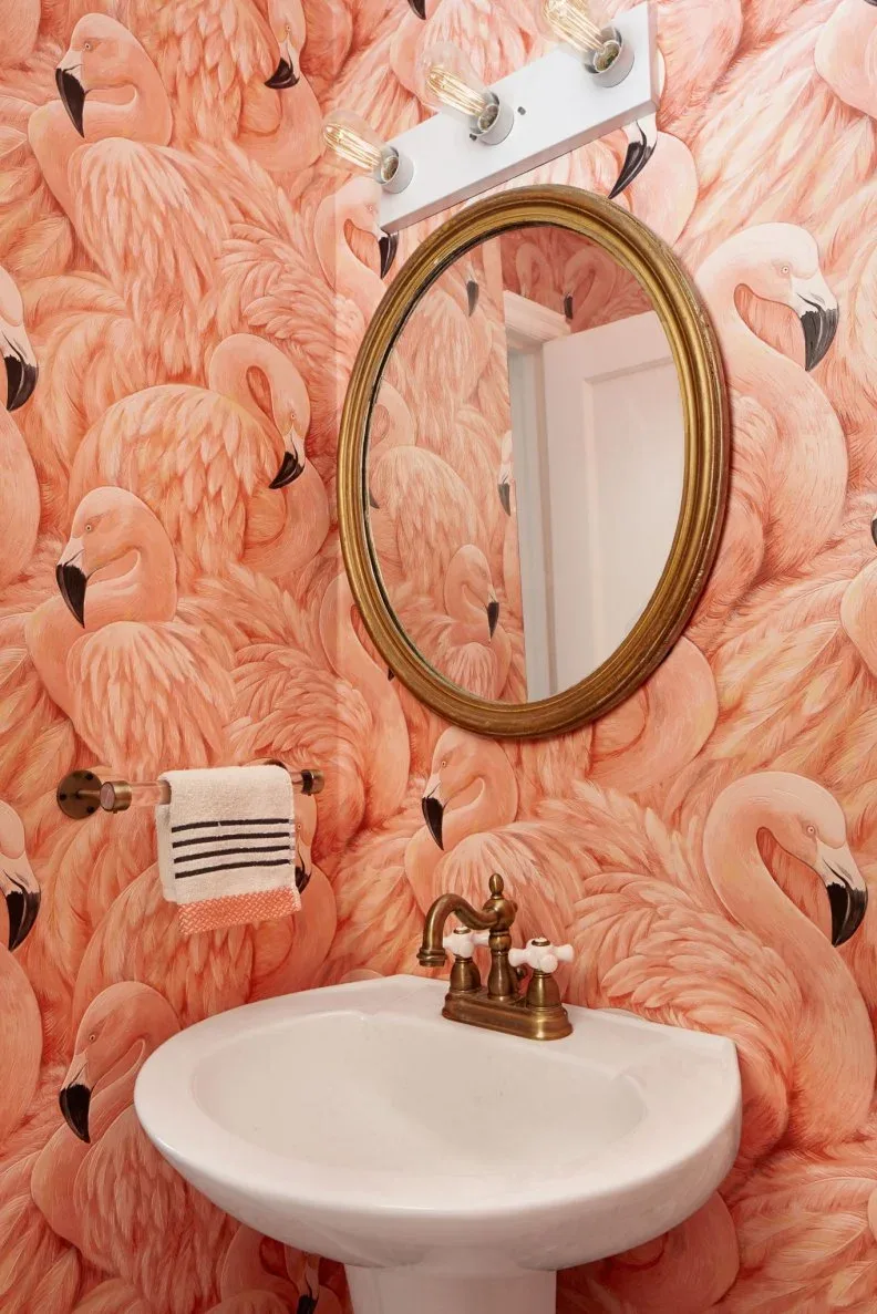 Powder room detail with pink flamingo wallpaper, oval golden-framed mirror, white pedestal basin, striped towel and a row of exposed-bulb lights.