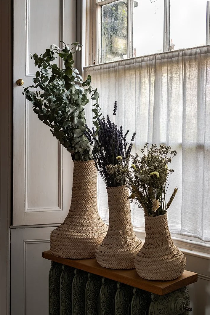 Three woven bottle-shaped vases of different heights hold leafy stems, purple flower spikes and lighter dried flowers beside a curtained window.