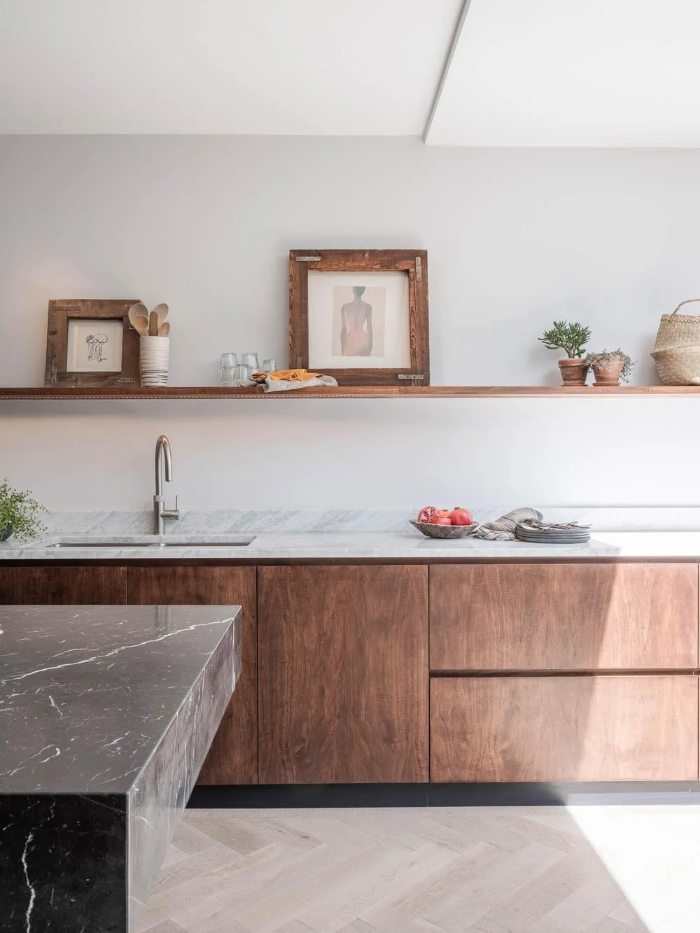 Handleless walnut base cupboards sit beneath a pale counter and a long wood shelf, with a stainless faucet and white wall.
