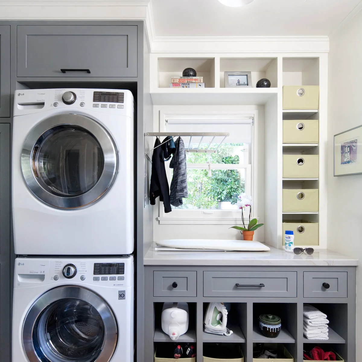 Gray-and-white laundry room with stacked appliances, a small ironing board on the counter and cubbies with pale green bins