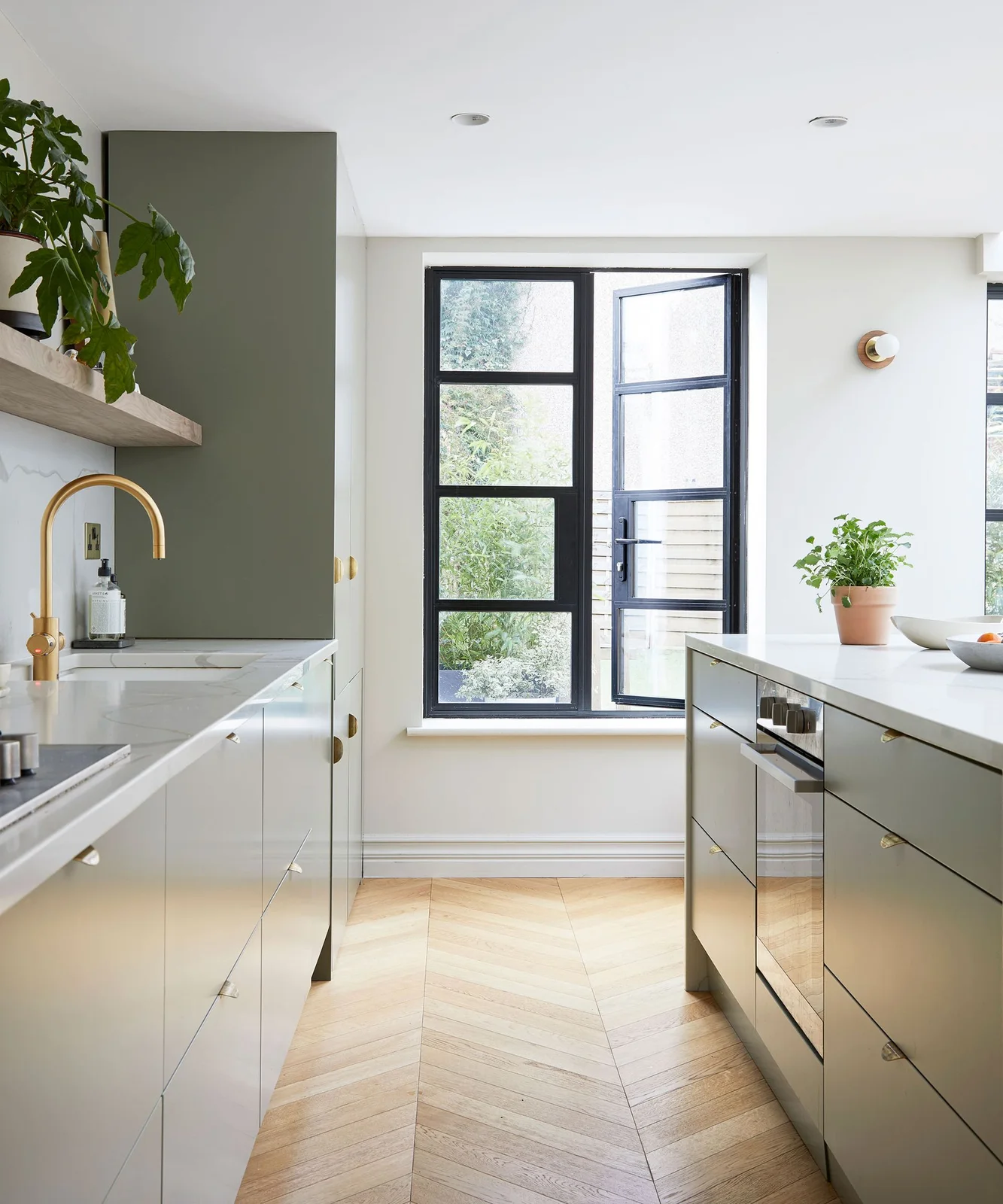 A galley kitchen has pale and gray-green cabinets, a gold-colored tap and a black-framed window