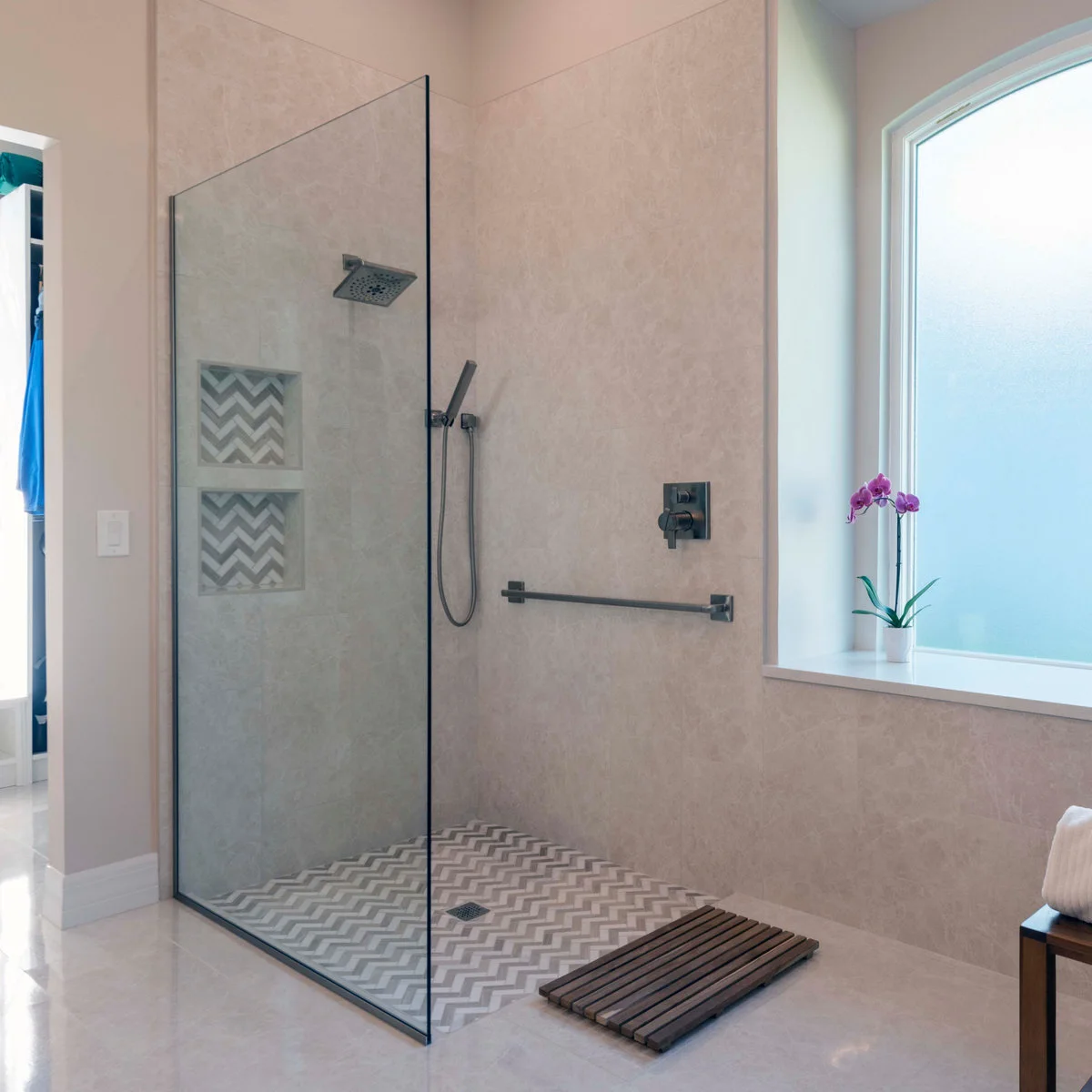 A fixed glass screen stands beside an open shower entrance with chevron floor tiles, two niches and a horizontal grab bar.