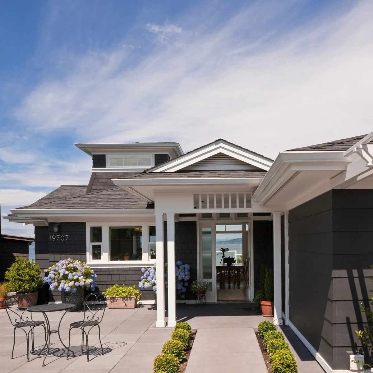 A dark gray house with a front sitting area, low bright-green shrubs beside the walk and blue-flowered pots.