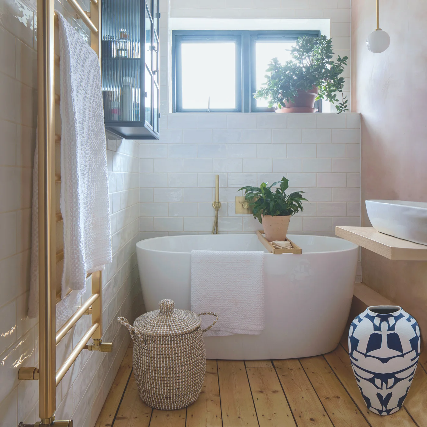 A freestanding white bath sits beside wooden floorboards, white wall tiles, pale pink walls, potted plants and a woven basket