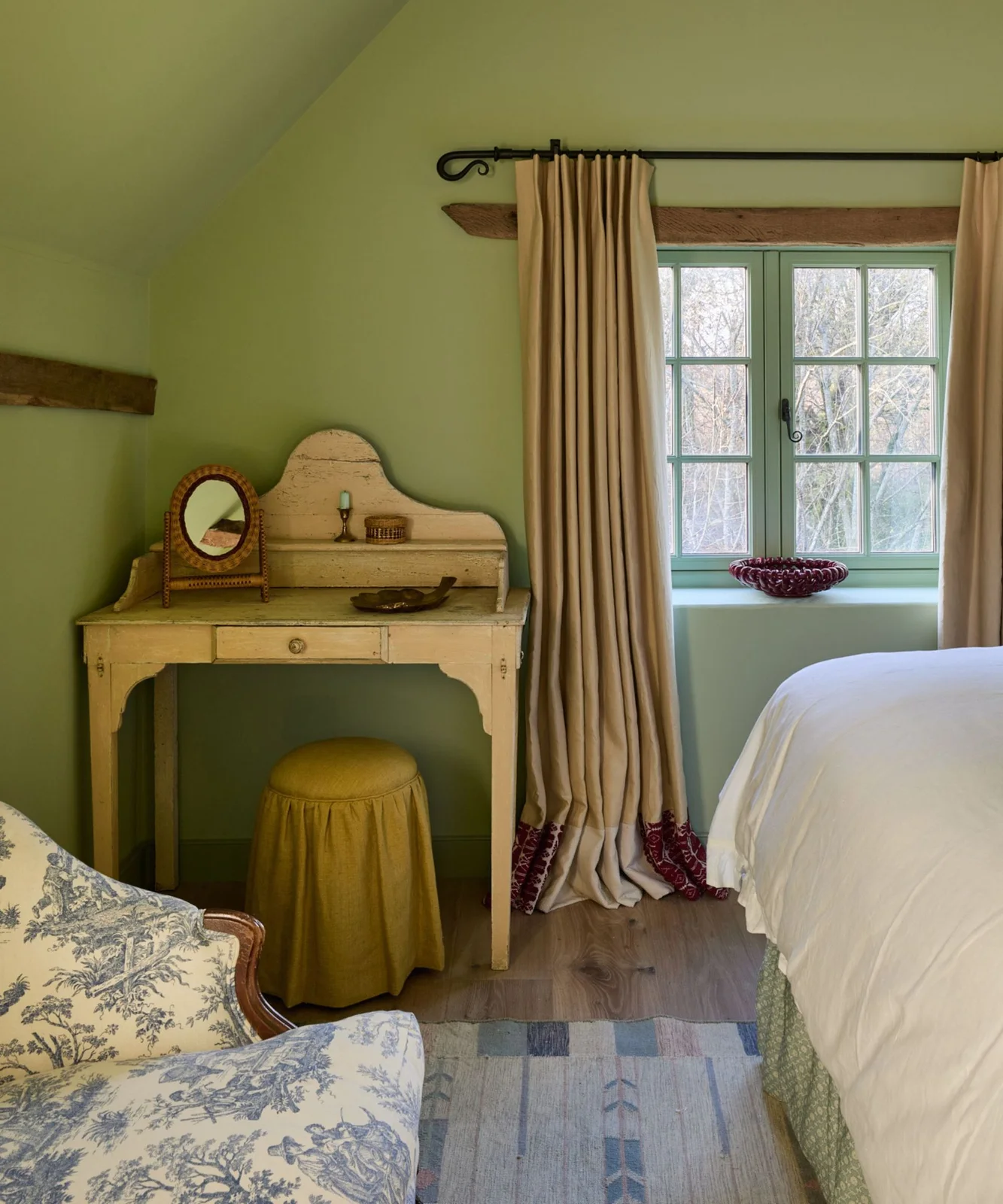 Soft green bedroom walls around an aged pale dressing table, beige curtains and a small ochre stool.