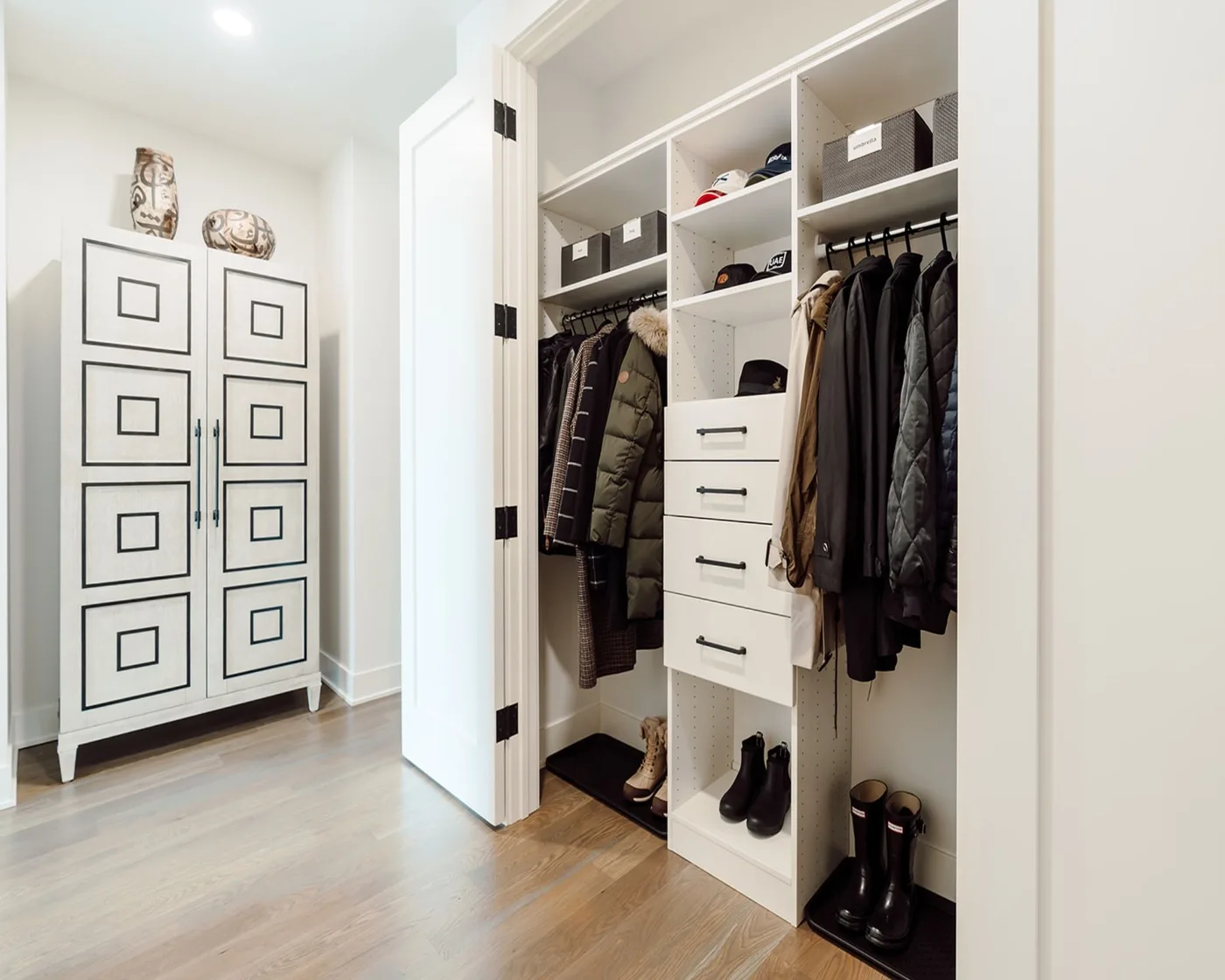 Open entry closet with folding doors, two coat rails, central drawers, upper labeled bins, shoe shelves and boots below