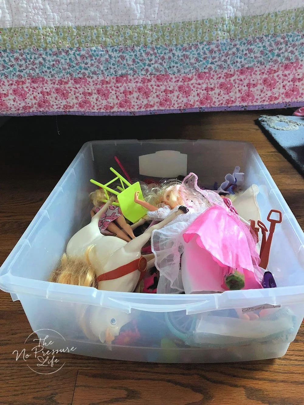 Wide clear bin in front of a bed containing toy horses, doll furniture and dressed dolls.
