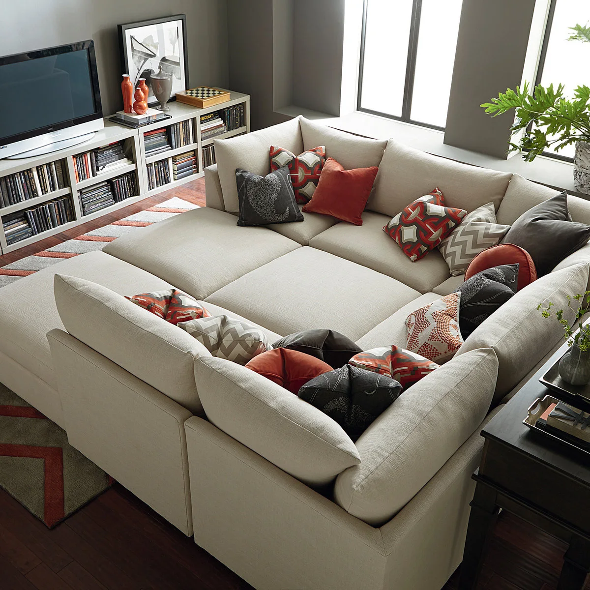A cream modular sofa with broad chaise sections faces low book storage and a television beside tall windows.