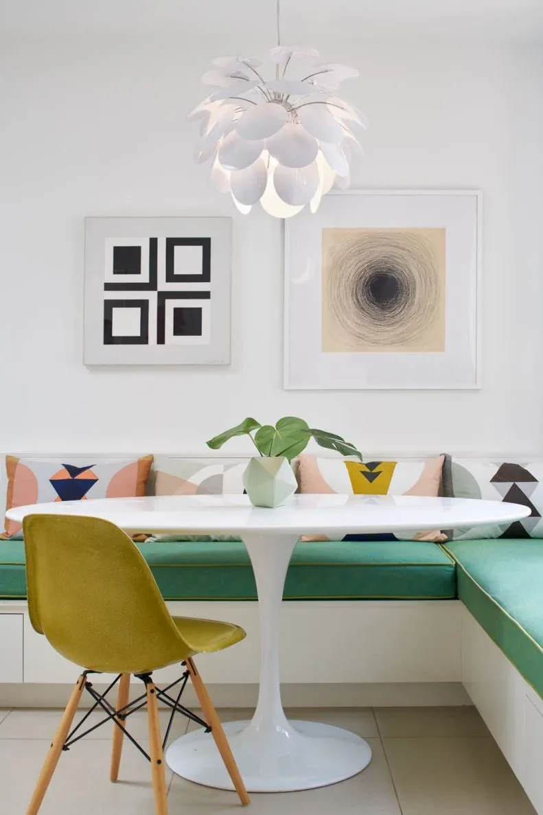 Two pale-framed graphic pictures above a green dining bench with geometric cushions and a white pedestal table