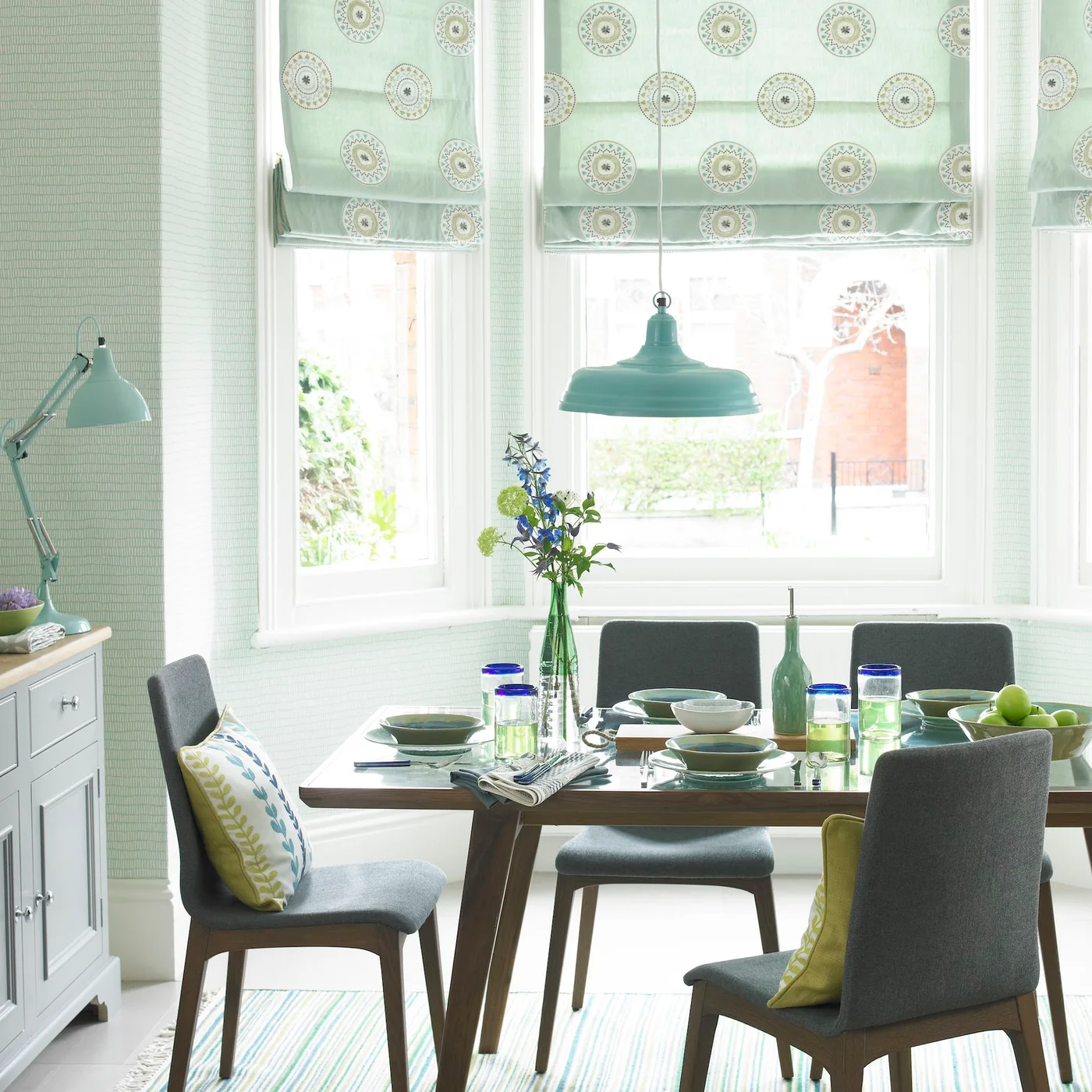 Patterned pale green blinds and green light fittings sit above a wooden table with gray chairs, pale sideboard and green glassware