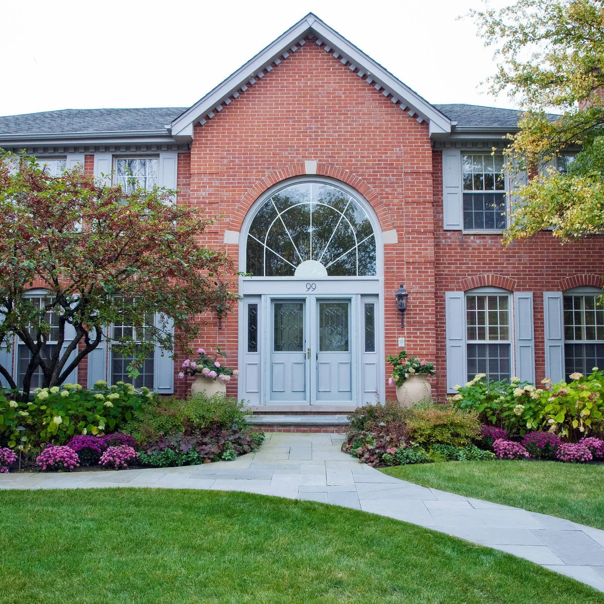 A pale paved front path divides flower beds with pink blooms and green shrubs beside a lawn.