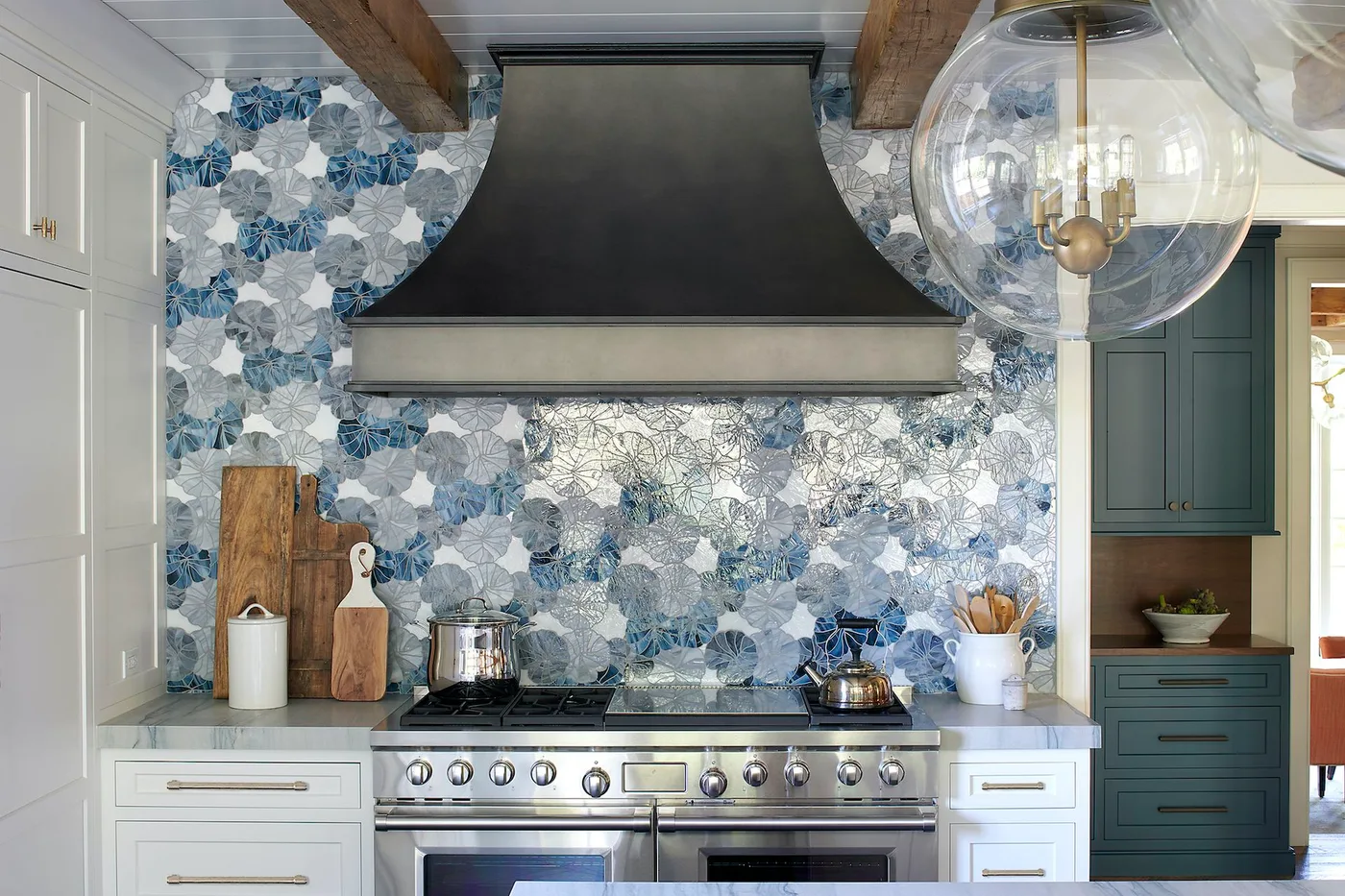 Country kitchen cooking wall with blue and gray floral pattern, dark broad hood, pale lower cabinets, exposed beams and round glass hanging lights.