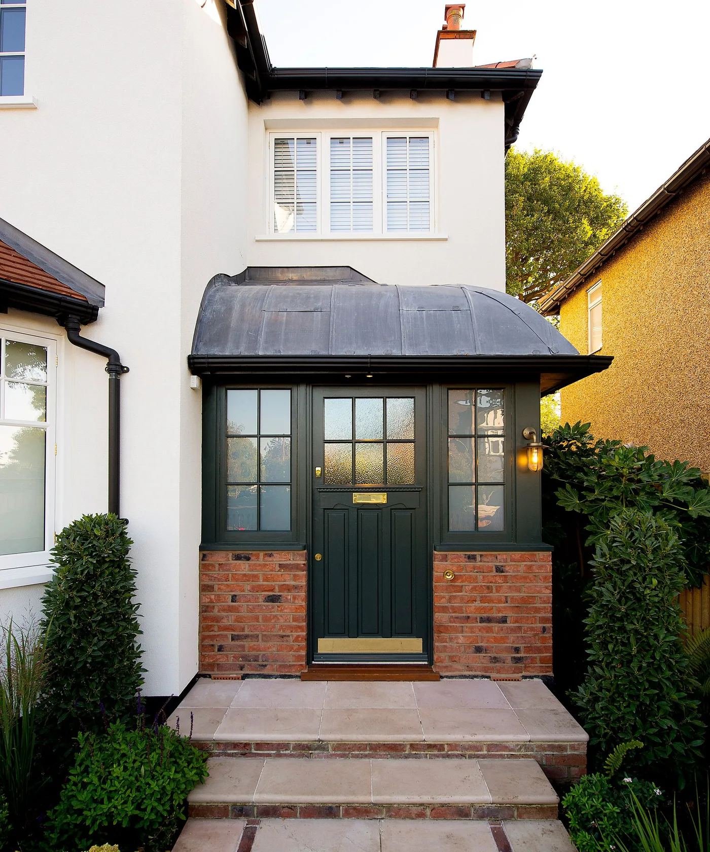 Small enclosed country porch with dark green glazed entrance, brick base, pale upper walls and shrubs