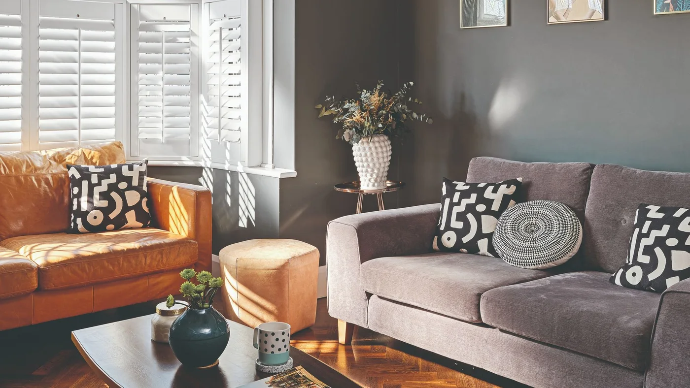 A gray sofa and warm brown sofa face a low table near white shutters against a dark gray wall with graphic cushions