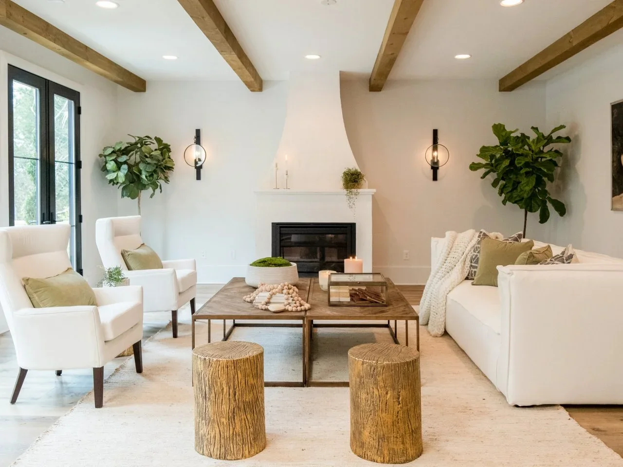 White chairs and a white sofa face paired timber coffee tables before a pale fireplace beneath exposed beams.