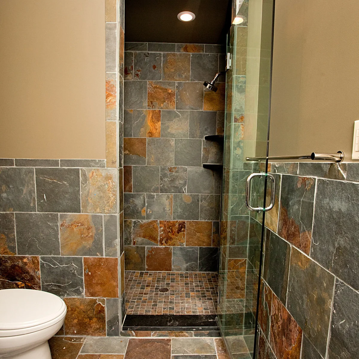 A narrow shower with gray and rust-colored tiles, dark corner shelves and a glass door, with matching tile on the bathroom walls.