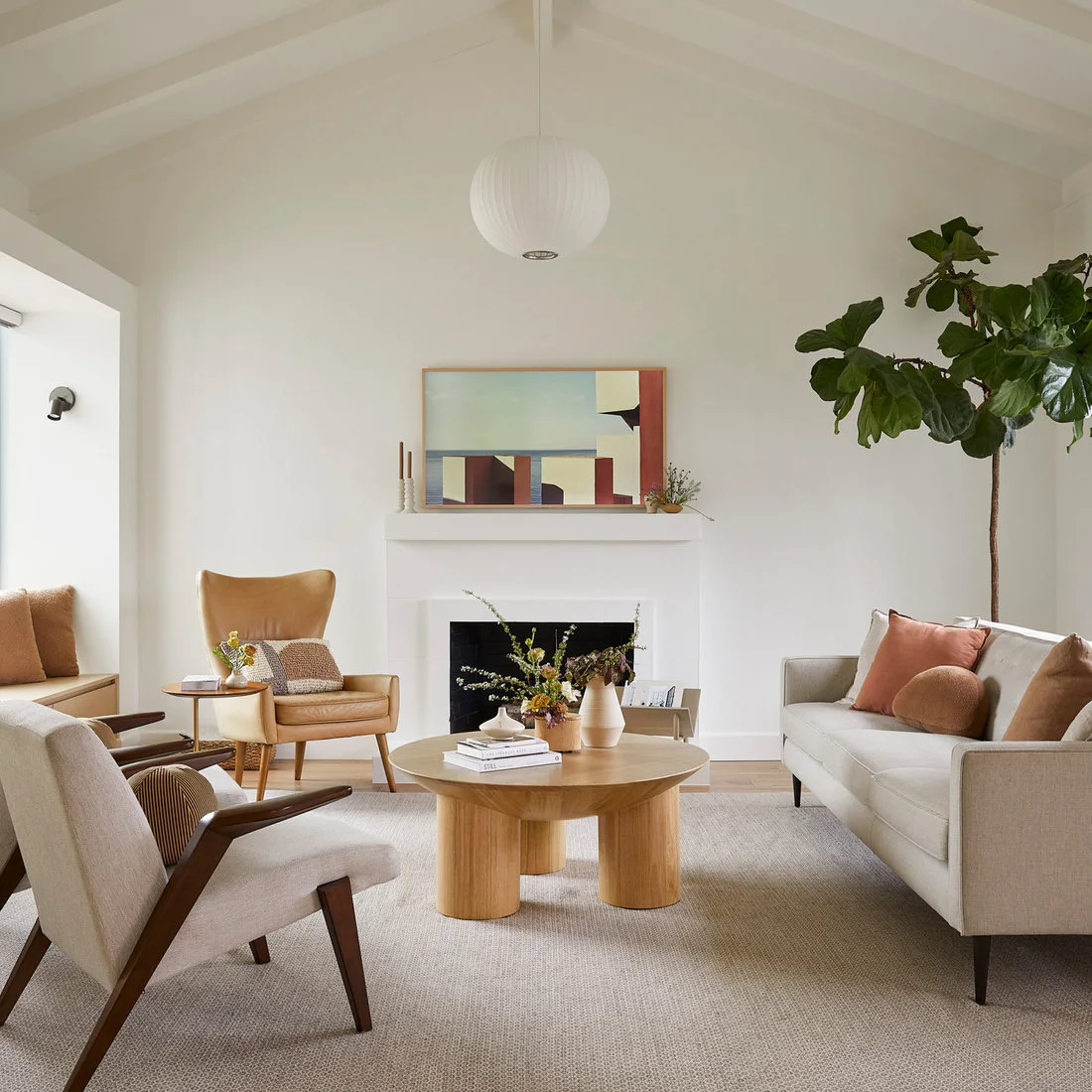 A pale sofa and wood-framed chair sit around a round wood table beside a tan chair, white fireplace and tall plant.