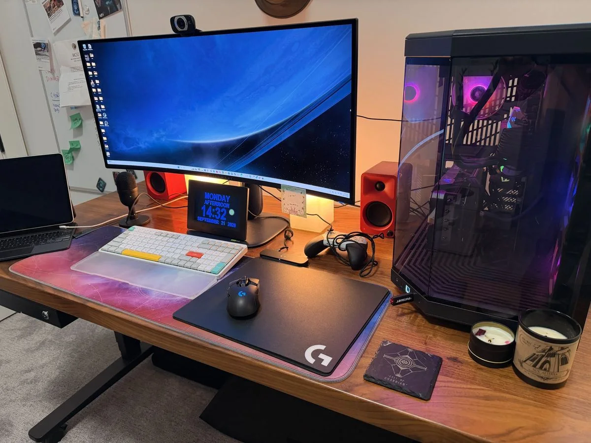 A wood desk with a large pink-and-blue mat, a white keyboard, red speakers, a single monitor and a glass-sided computer tower.