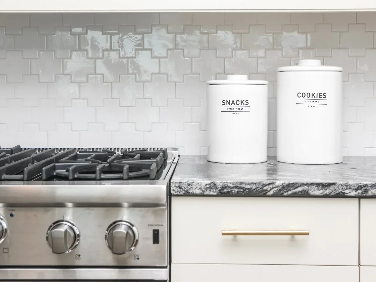 Glossy cross-shaped white kitchen tiles form a backsplash beside a range, pale drawers and a dark veined counter.
