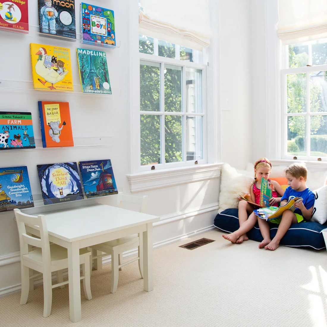 Children read on blue floor cushions beside bright windows, a small white table and chairs, and narrow wall ledges displaying colorful book covers.