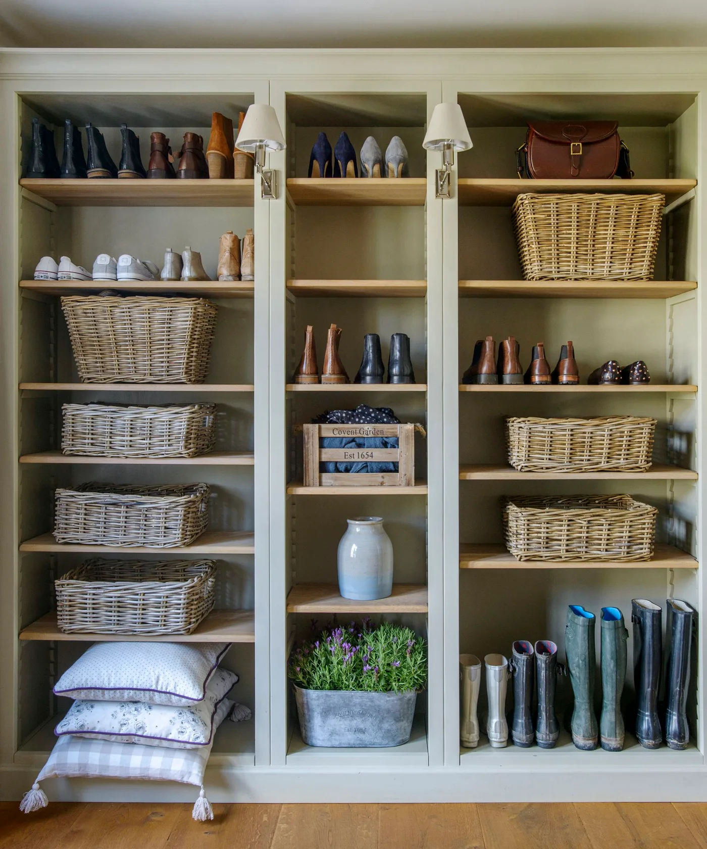 Green open mudroom shelving with shoes, tall boots, woven baskets and two small wall lights