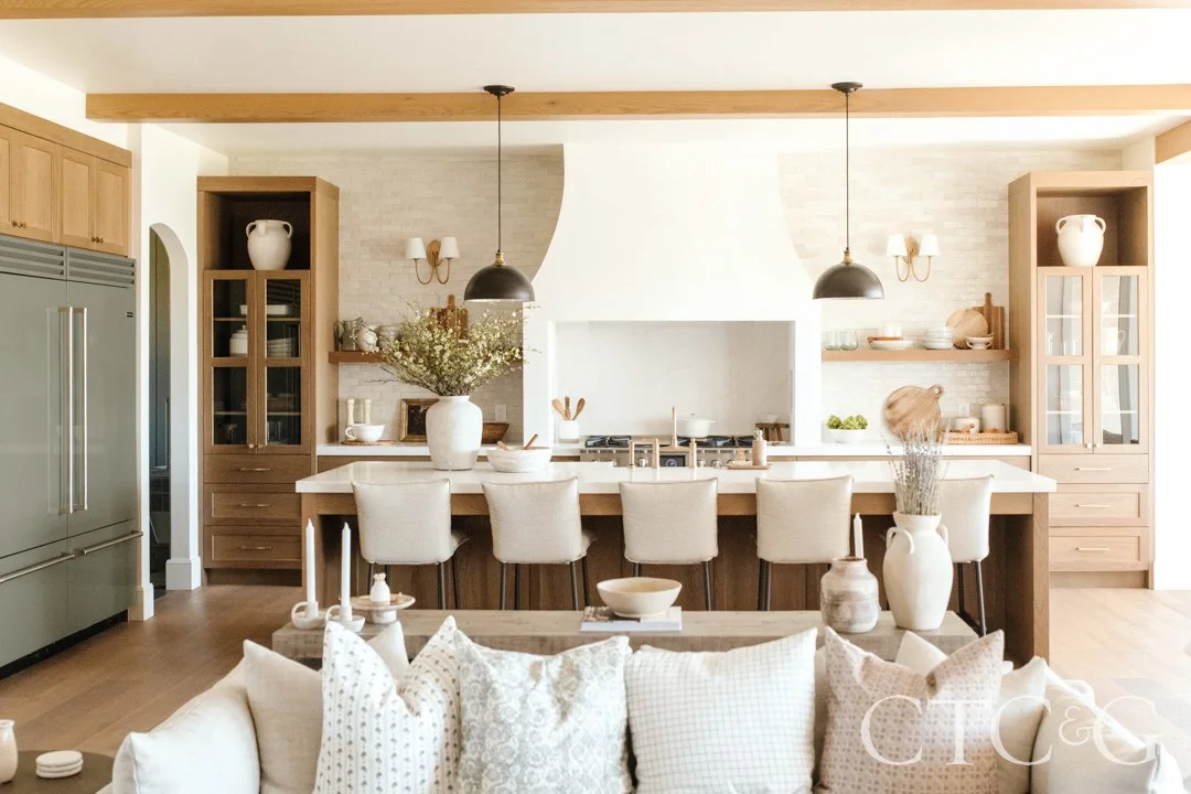 White oak cabinets and glass-front towers frame a white hood, with muted green appliances, open shelves and pale island stools.