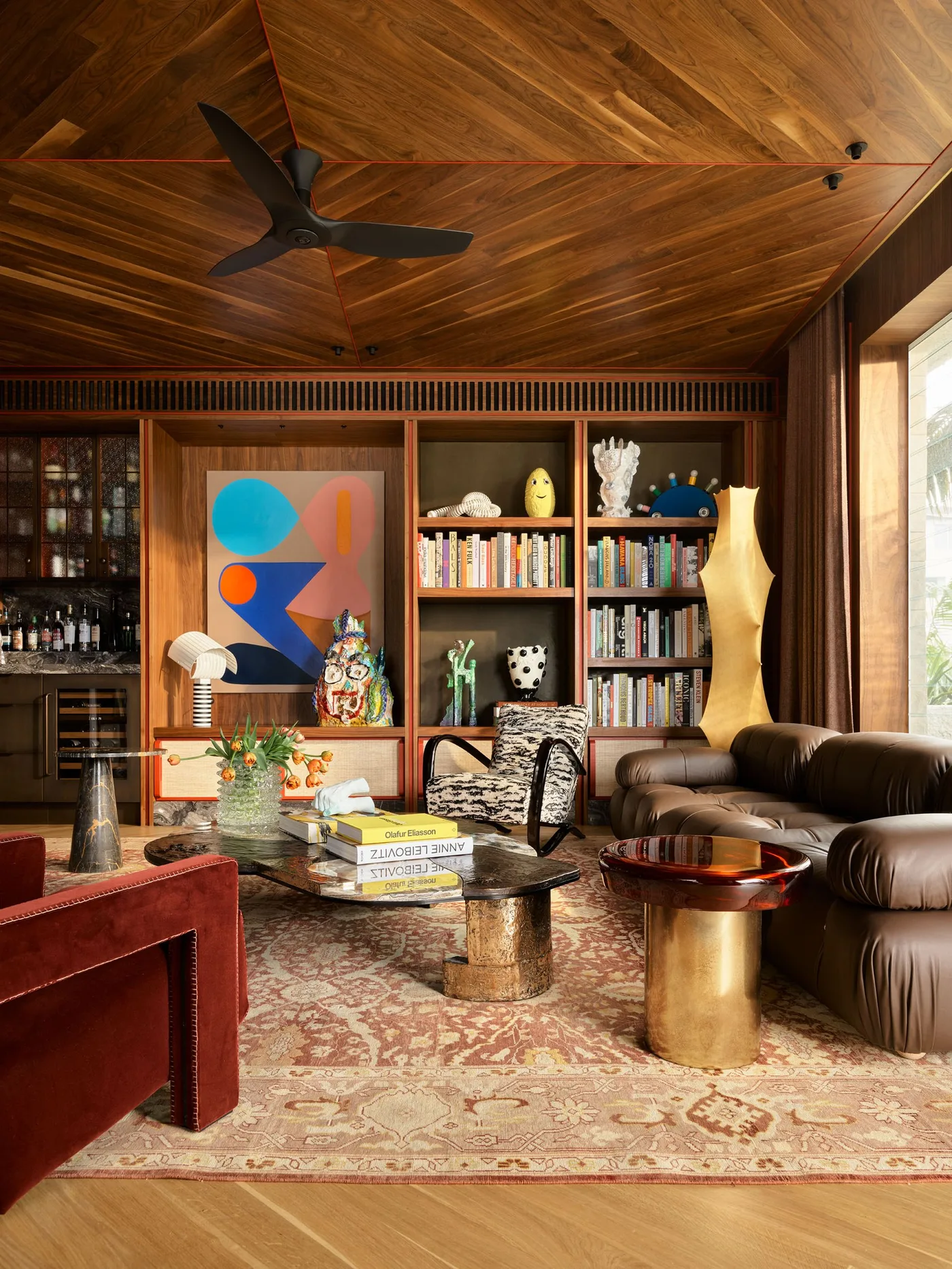 A low brown leather modular couch sits below a wooden ceiling beside bookshelves, a patterned rug and rounded metallic tables