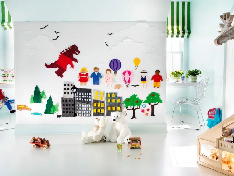 A bright white play area has a large felt scene with colorful people, trees and buildings, green striped window shades and a wood dollhouse at one side.