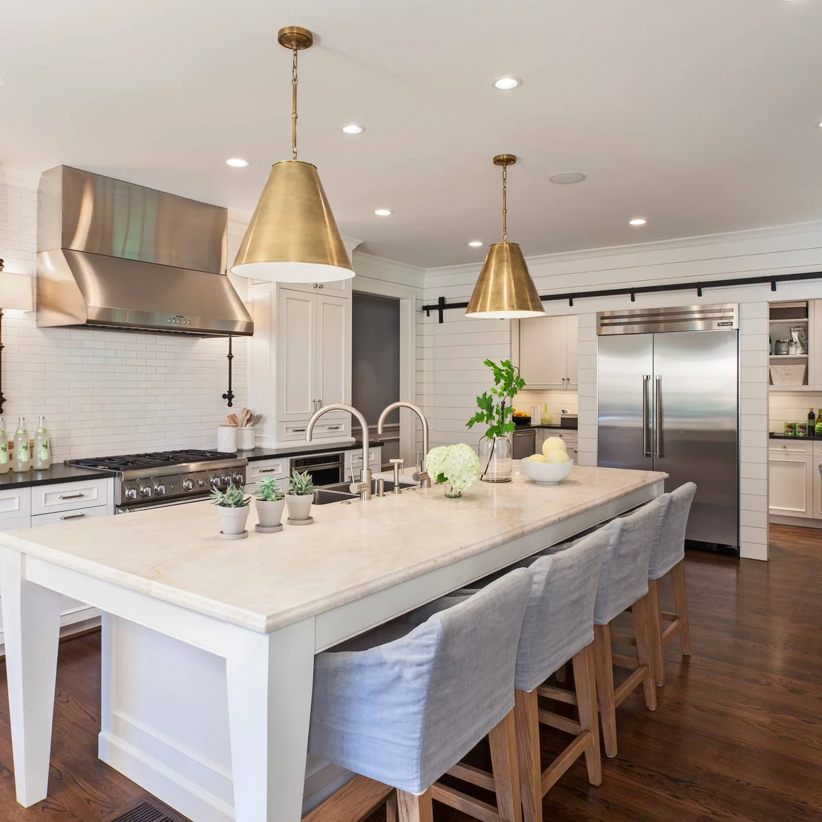 Two broad warm metal cone pendants hang over a pale island with gray upholstered stools and recessed ceiling lights.