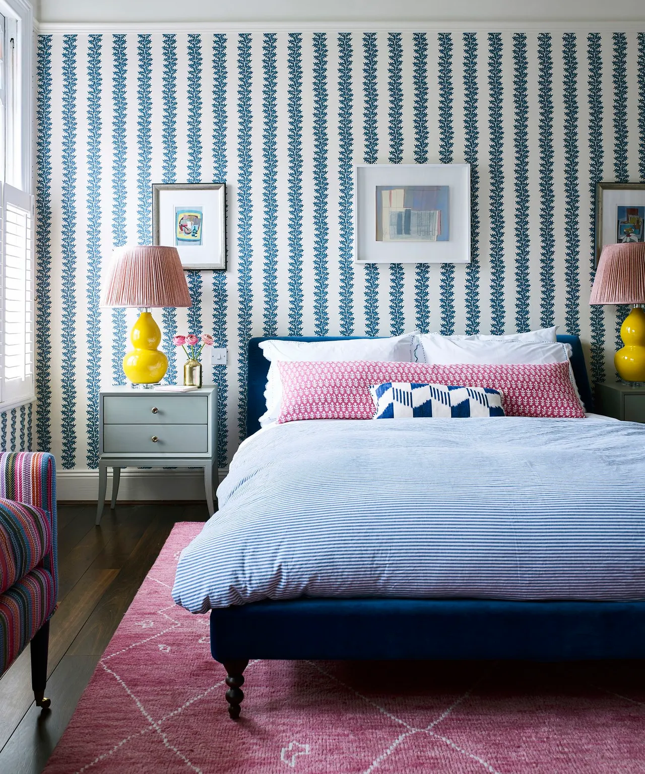 Vertical blue botanical bands rise behind a dark blue bed with pale bedding, pink accents and yellow bedside lamps.