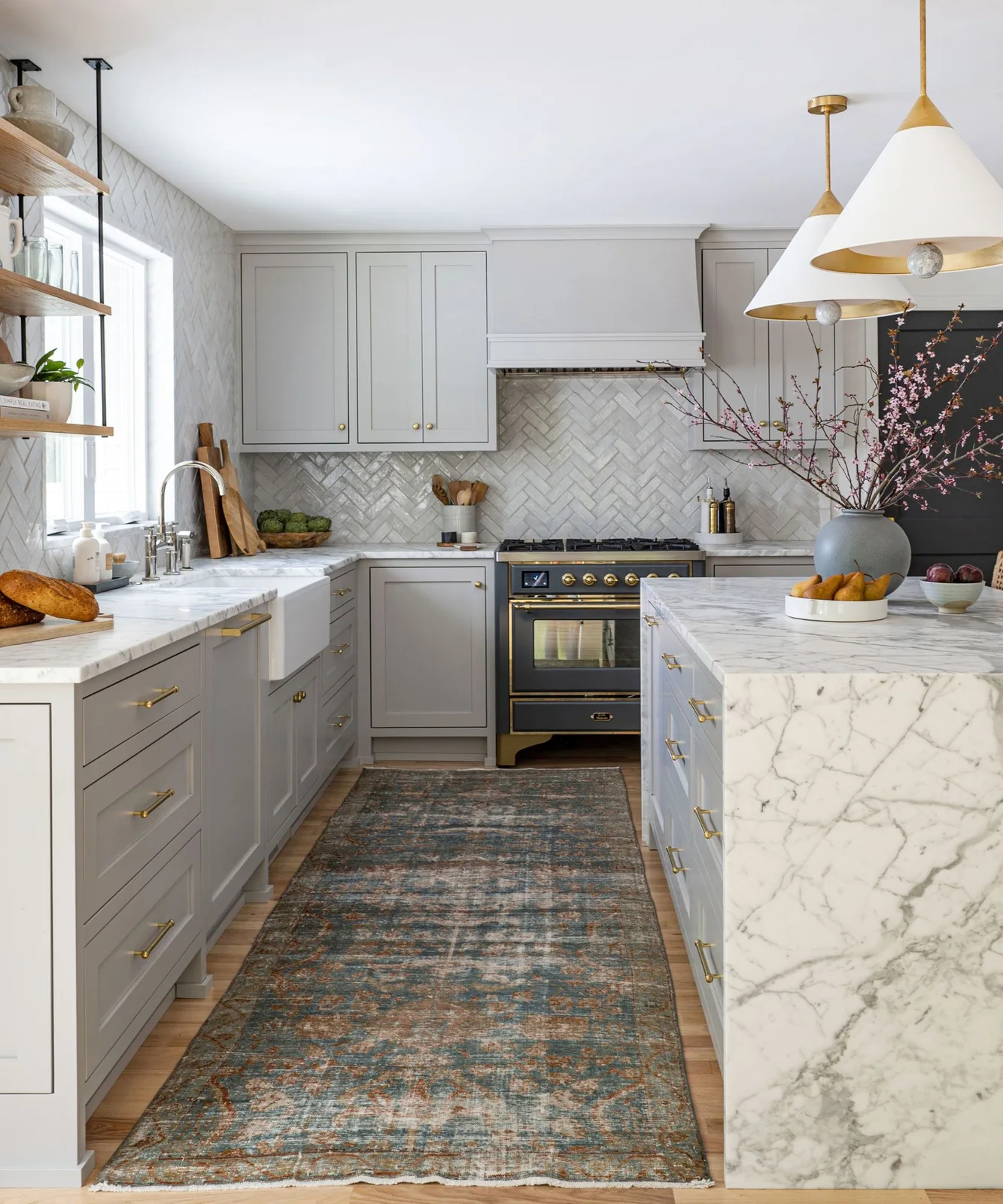 Pale gray cabinets with gold-toned pulls surround a strongly veined island and herringbone backsplash beside a dark range cooker