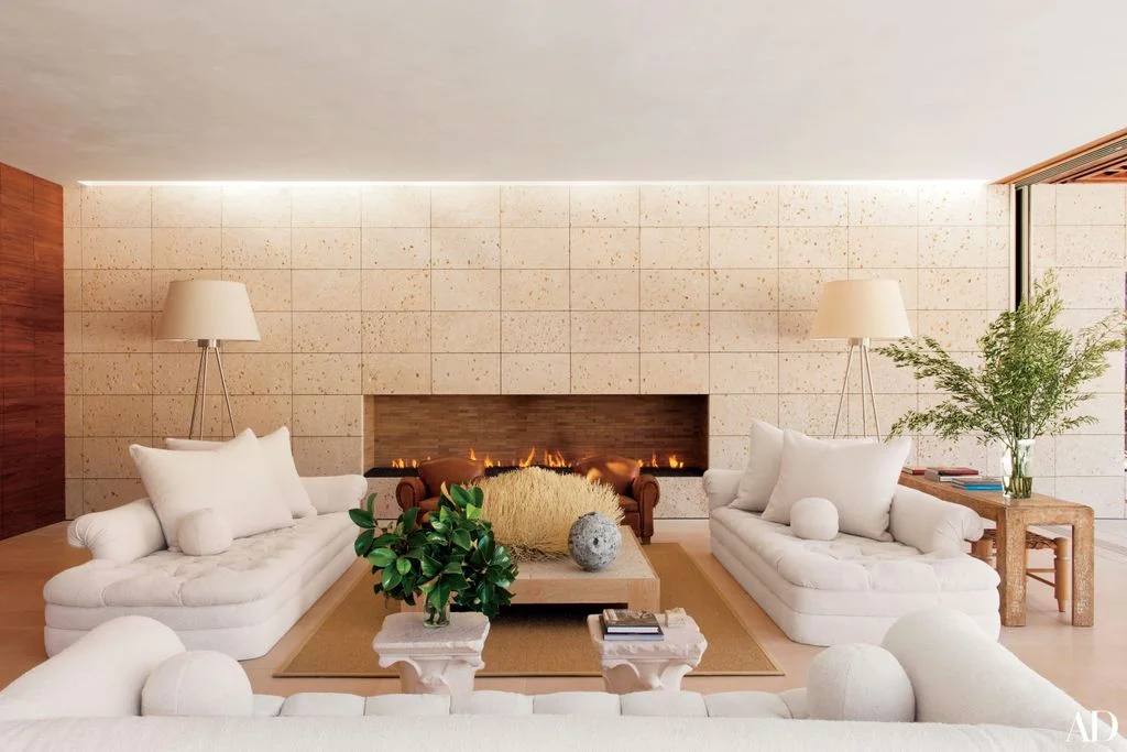 A long horizontal fireplace opening sits within a pale rectangular wall grid behind white sofas and warm wood tables beside two shaded floor lamps
