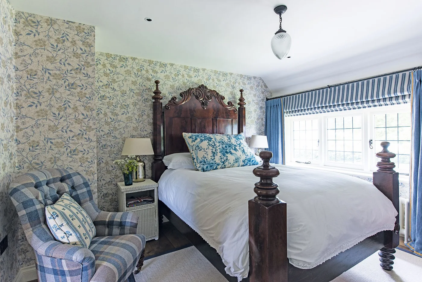 Bedroom with a dark carved wooden bed, blue floral wallpaper, a checked armchair and a striped window valance.