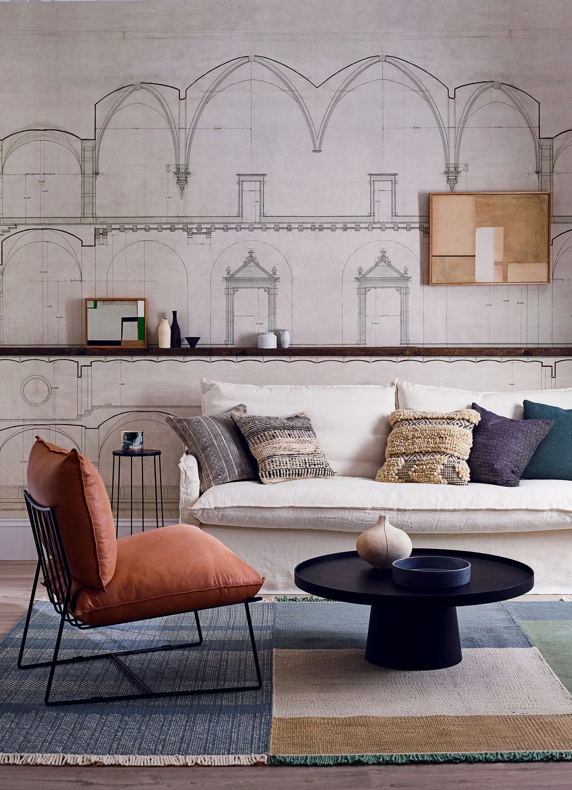 An architectural drawing wallpaper fills the wall behind a white sofa, a picture ledge and an orange chair.