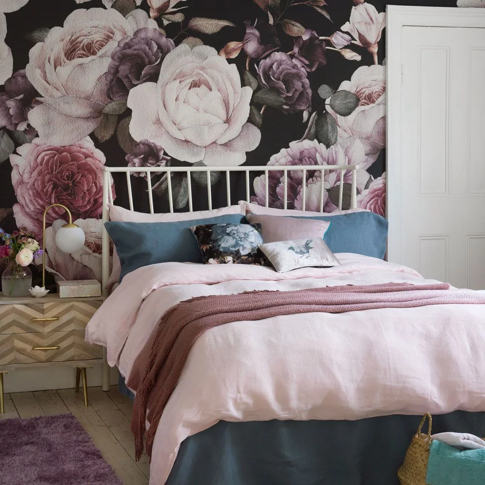 An oversized pink rose wallpaper covers a black bedroom wall behind a white metal bed and pink bedding.